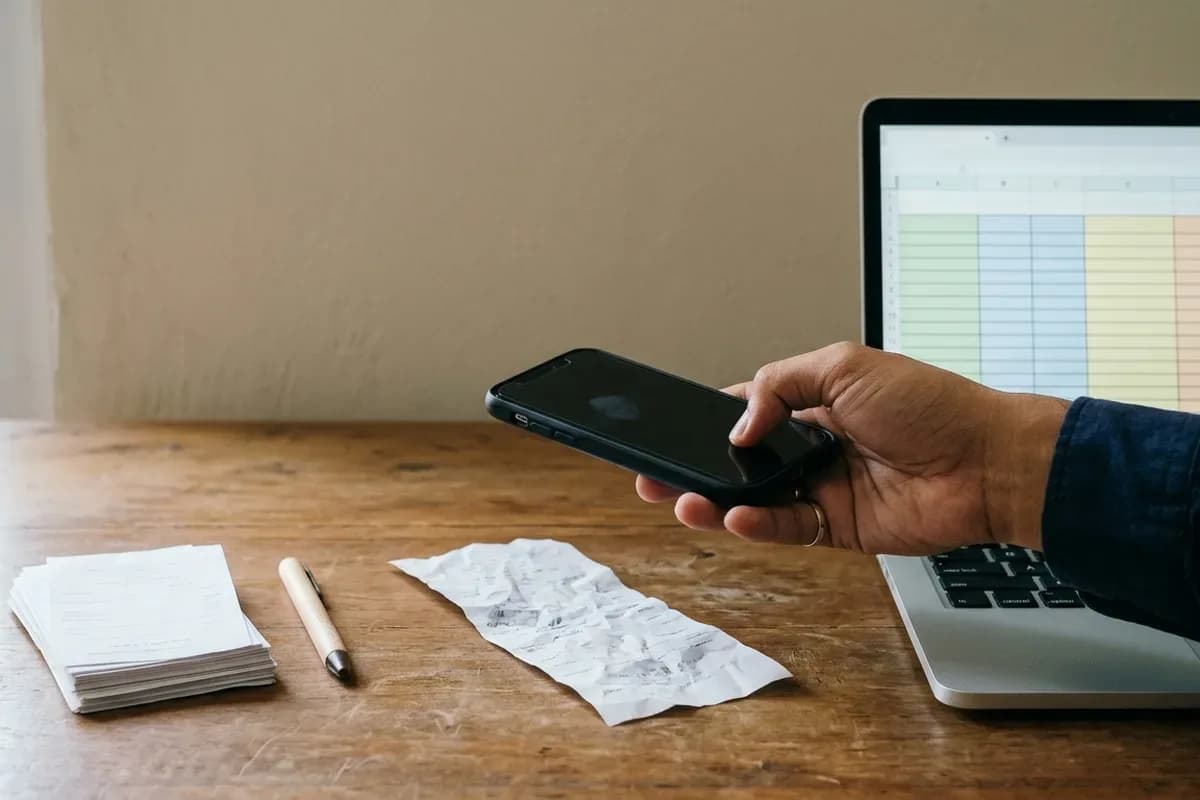 Tangan memegang ponsel untuk memotret nota belanja di meja kayu, di samping tumpukan nota dan laptop yang menampilkan spreadsheet kosong